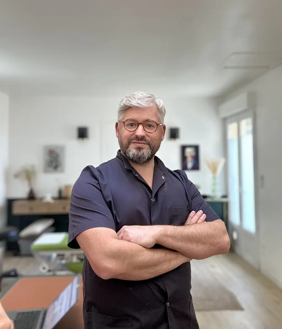Olivier, bras croisés, en tunique bleu marine, dans la salle de soin